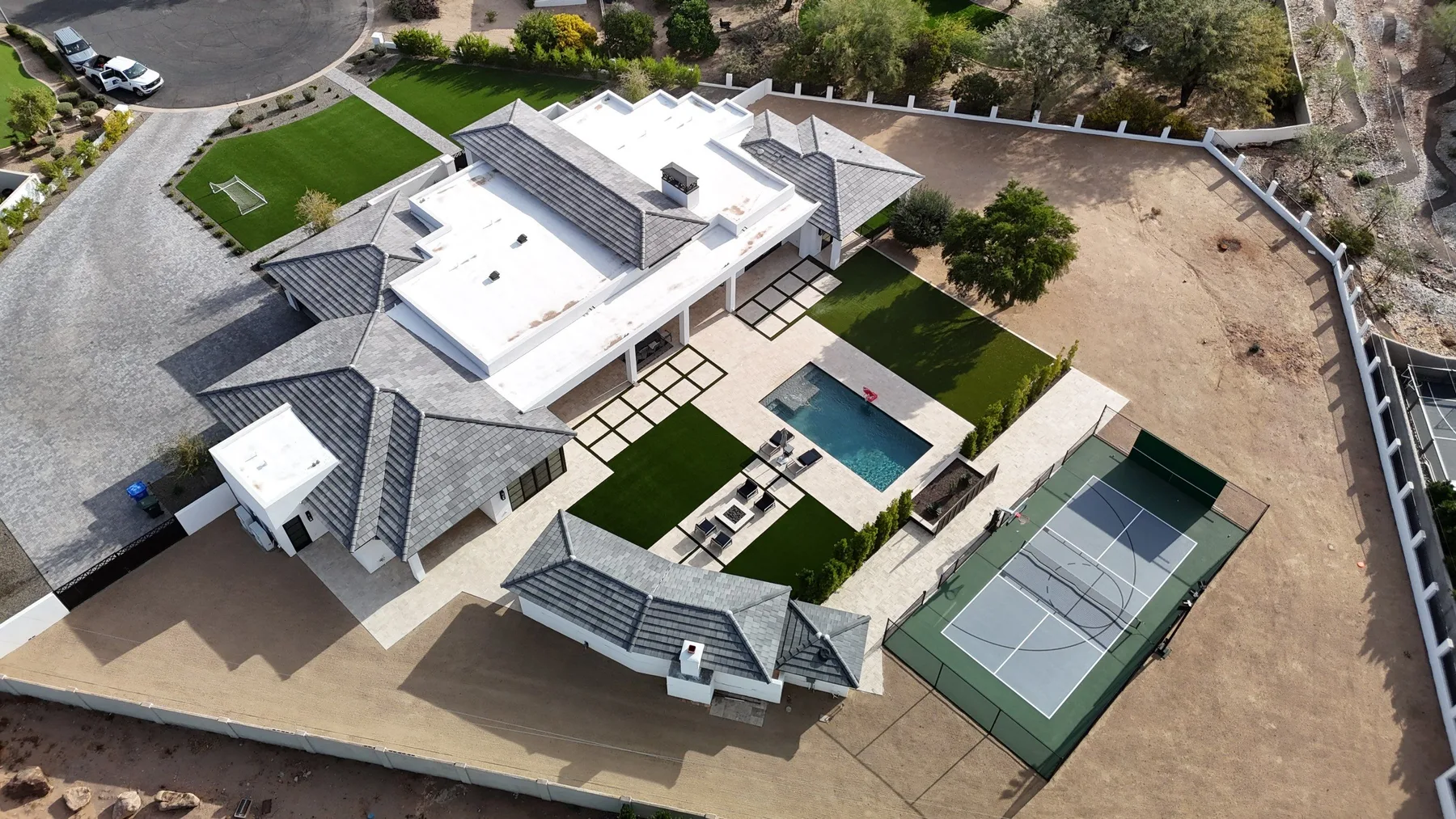 High oblique aerial view of white flat roof sections, gray tiled wings, pool, and court