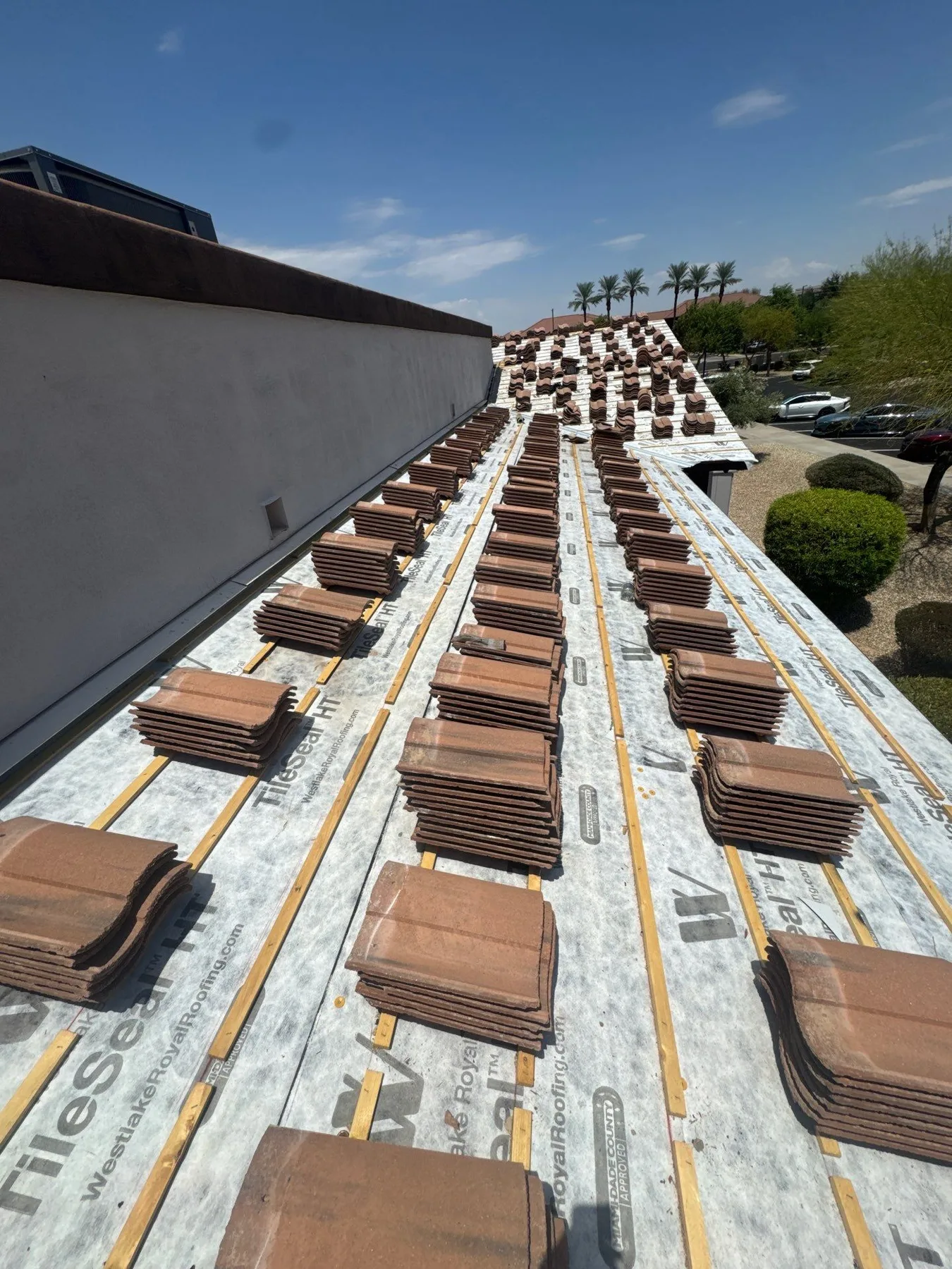 Close rooftop view of white underlayment and spaced brown tile bundles beside a parapet
