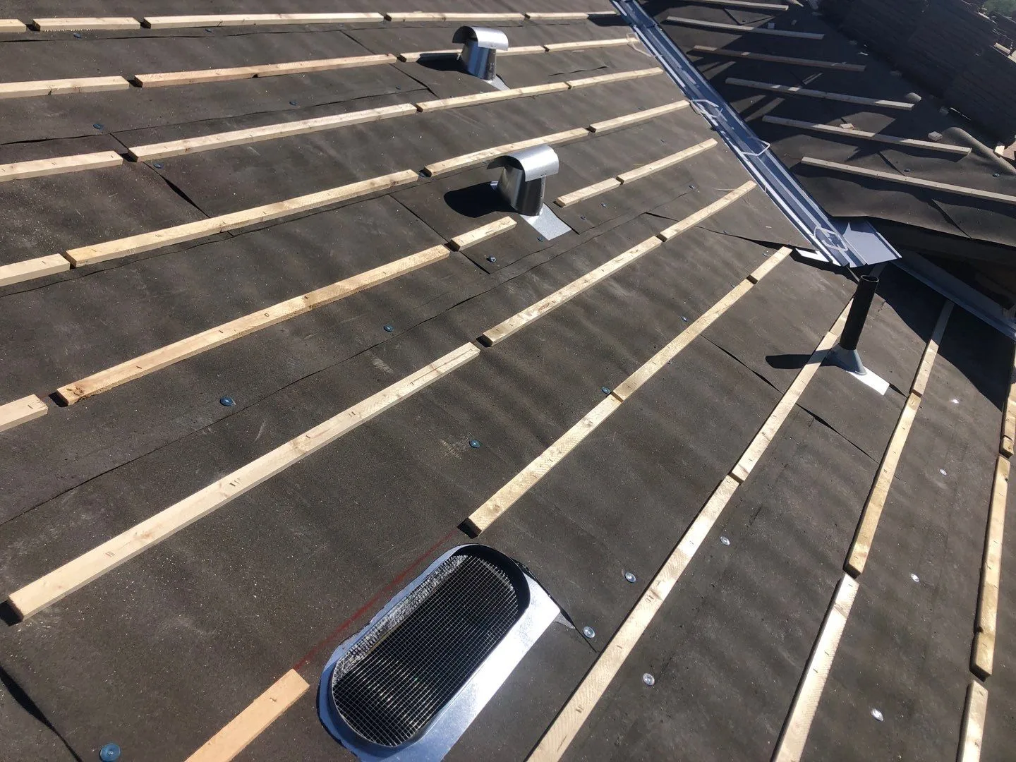 Close rooftop view of black underlayment, pale battens, vent pipes, and metal valley flashing