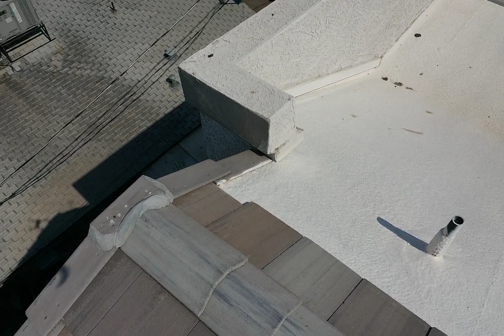 Close overhead view of white coated flat roofing beside gray shingles, parapet, and vent pipe