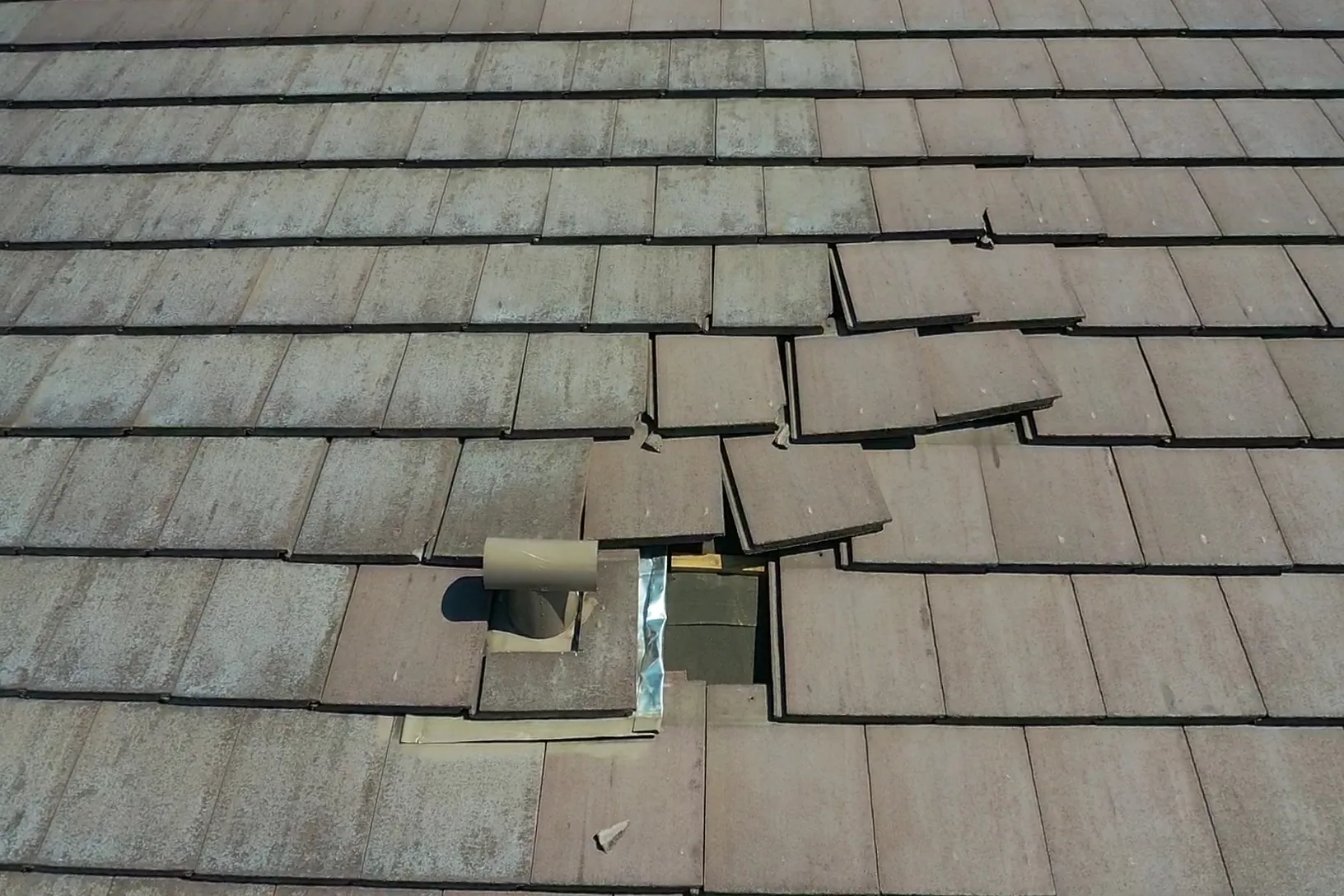 Close overhead view of weathered gray tiles with displaced pieces surrounding a pipe flashing