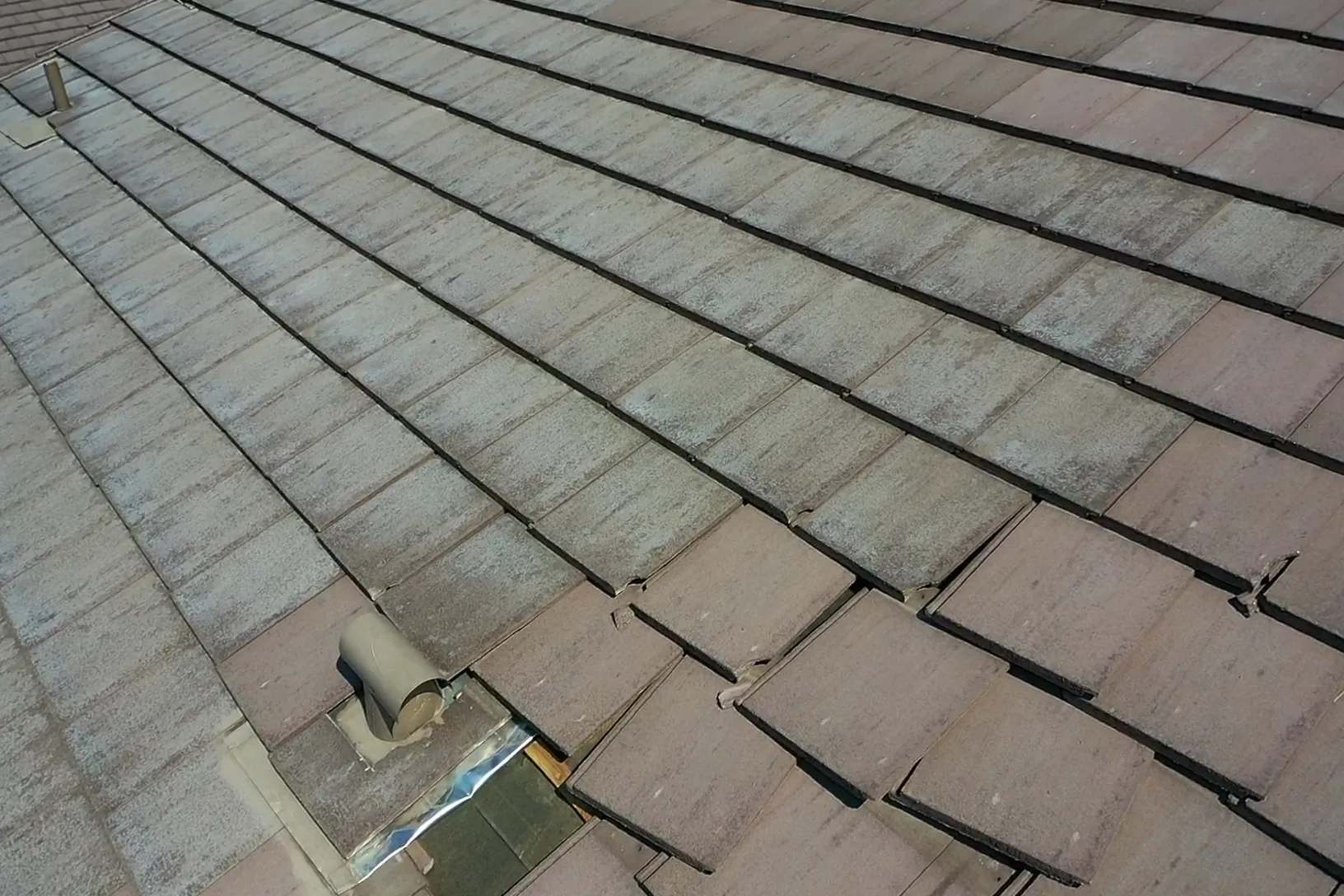 Angled overhead view of weathered brown-gray flat tiles, open gaps, and metal pipe flashing